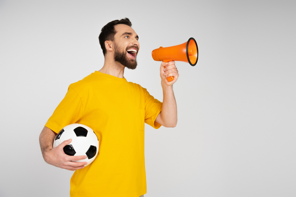 fröhlicher bärtiger Mann schreit in Lautsprecher, während er den Fußballball isoliert auf grau hält - Foto, Bild