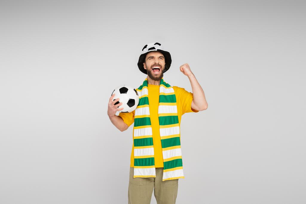Aufgeregter Sportfan mit Hut und Schal hält Fußballball in der Hand und zeigt Triumphgeste isoliert auf Grau - Foto, Bild