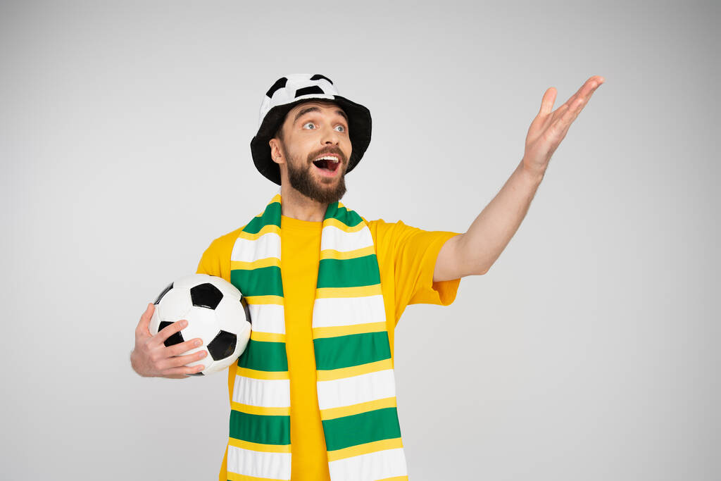 užaslý a radostný sportovní fanoušek v klobouku a šále držící fotbalový míč a ukazující rukou izolovanou na šedou - Fotografie, Obrázek