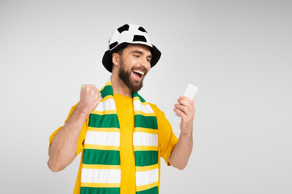 サッカーファンの帽子とストライプのスカーフで興奮した男が勝つジェスチャーを示しながら、グレーで隔離されたスマートフォンで試合を見て - 写真・画像