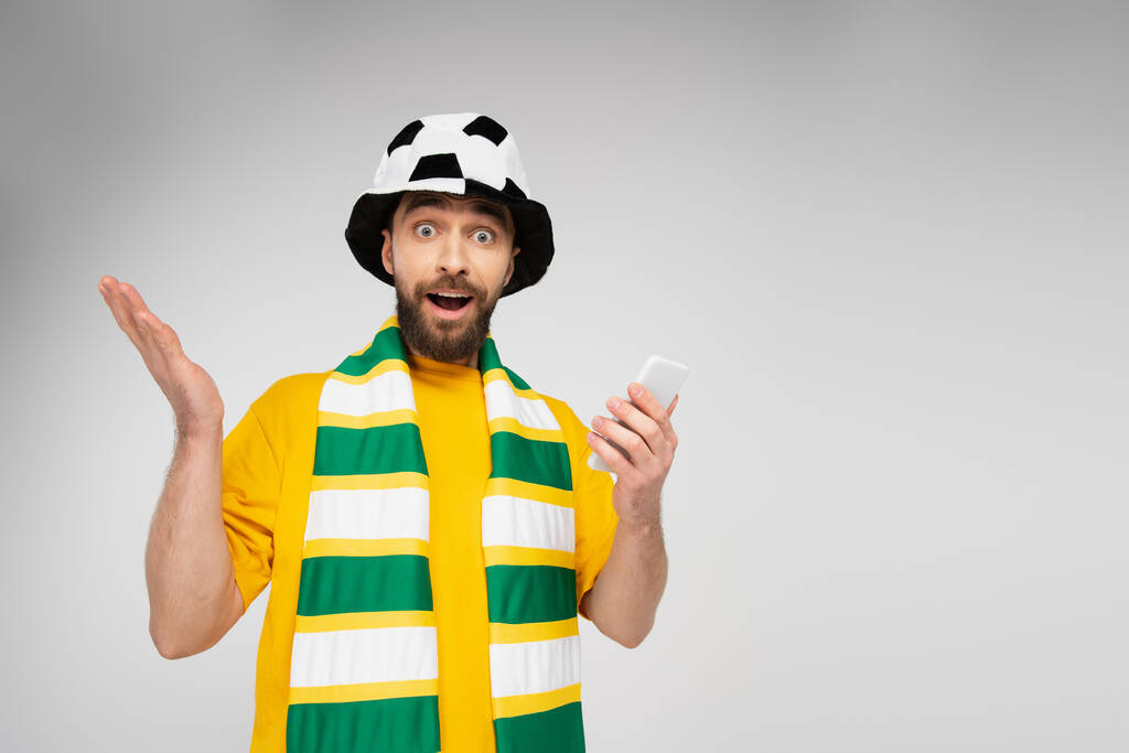 έκπληκτος άνδρας με ριγέ κασκόλ και καπέλο οπαδού ποδοσφαίρου κρατώντας κινητό τηλέφωνο και κοιτάζοντας κάμερα απομονωμένη σε γκρι - Φωτογραφία, εικόνα
