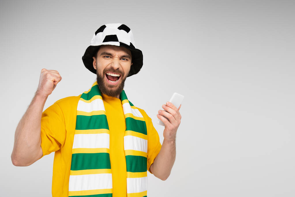 hombre alegre en el sombrero del ventilador de fútbol y bufanda rayada celebración de teléfono inteligente y regocijo aislado en gris - Foto, imagen