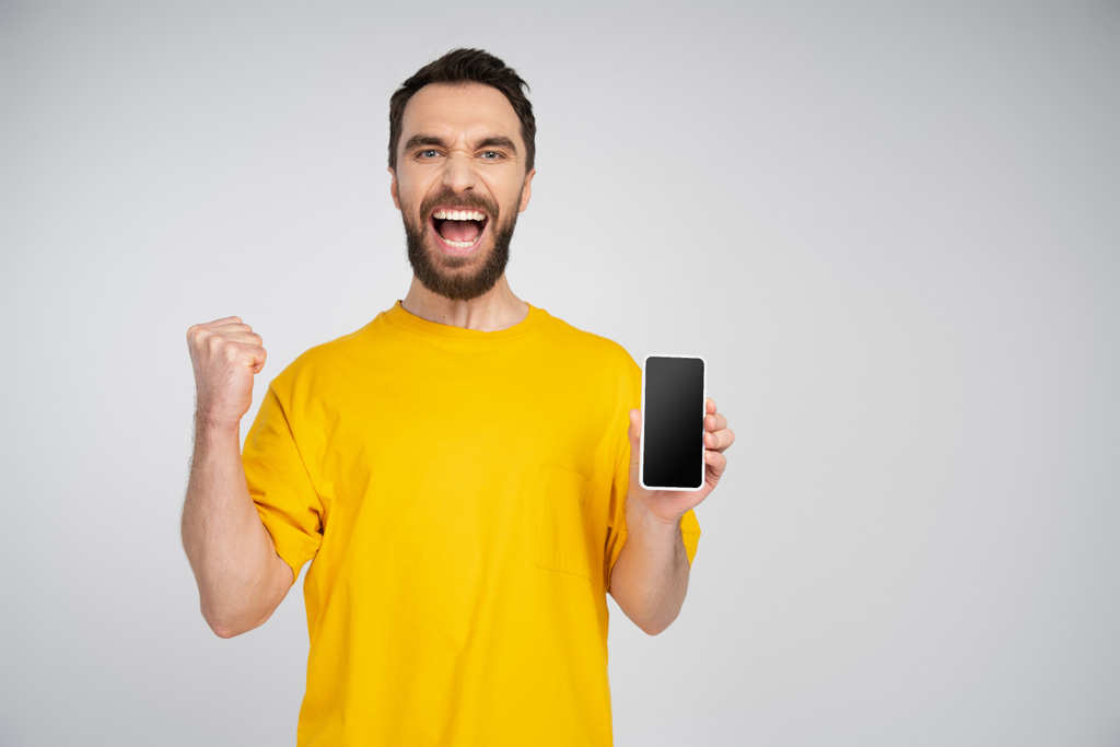 vzrušený vousatý muž držící smartphone s prázdnou obrazovkou a ukazující triumfální gesto při křiku izolované na šedé - Fotografie, Obrázek