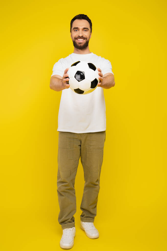 full length of happy bearded man in white t-shirt and beige pants holding soccer ball in outstretched hands on yellow background - Photo, Image
