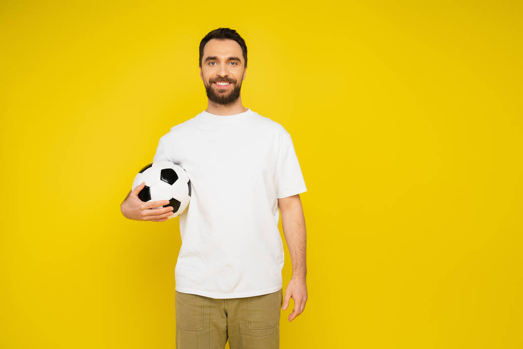Beyaz tişörtlü mutlu sakallı adam elinde futbol topuyla dikilip sarı renkli kameraya bakıyor. - Fotoğraf, Görsel