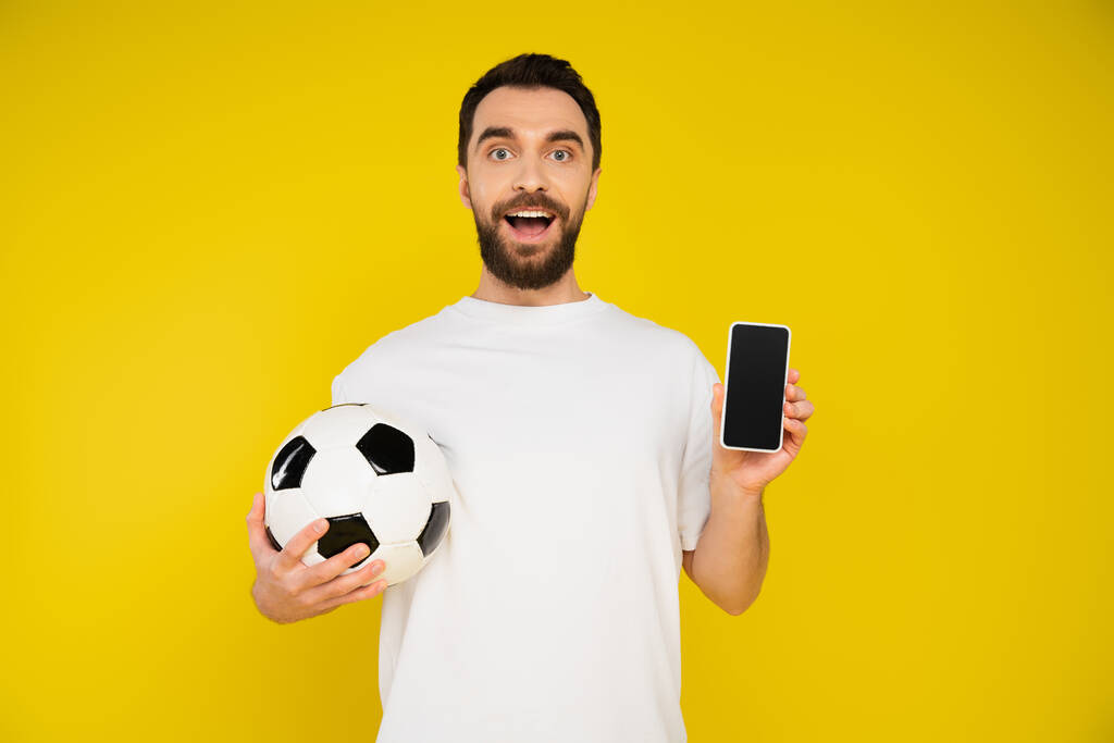 verbaasd voetbal fan holding bal en smartphone met blanco scherm geïsoleerd op geel - Foto, afbeelding