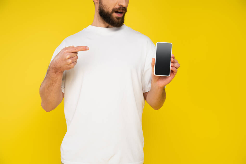 vista parcial del hombre barbudo en camiseta blanca apuntando al teléfono inteligente con pantalla en blanco aislado en amarillo - Foto, Imagen