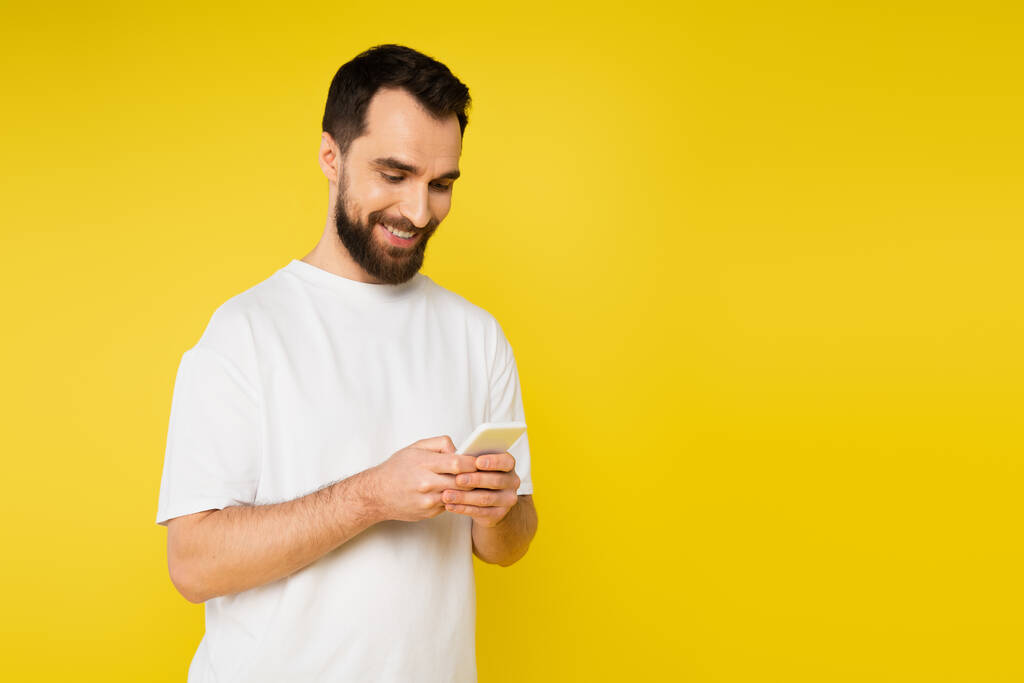 lachende brunette man met baard chatten op mobiele telefoon geïsoleerd op geel - Foto, afbeelding