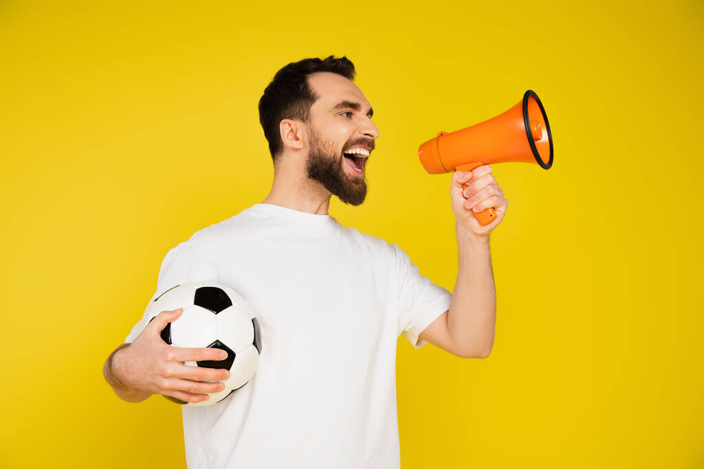 brodaty fan sportu w białym t-shirt trzymając piłkę nożną i krzycząc w megafon izolowany na żółto - Zdjęcie, obraz