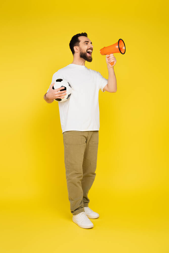 pełna długość człowieka w białym t-shirt i beżowe spodnie trzymając piłkę nożną i krzycząc w megafon na żółtym tle - Zdjęcie, obraz