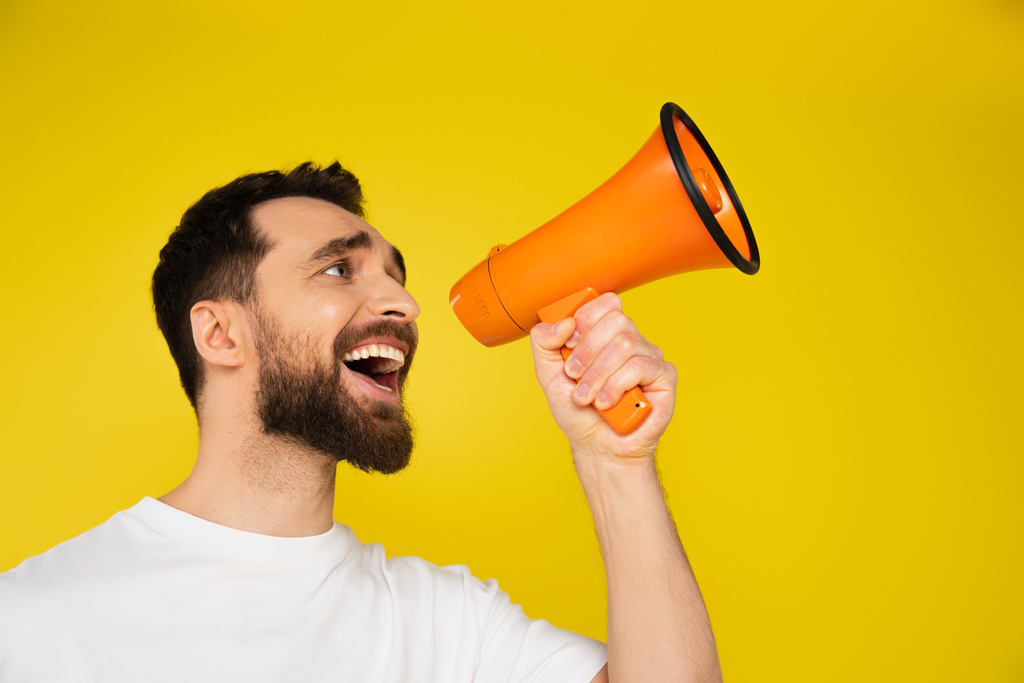 brunette bearded man shouting in megaphone while looking away isolated on yellow - Photo, Image