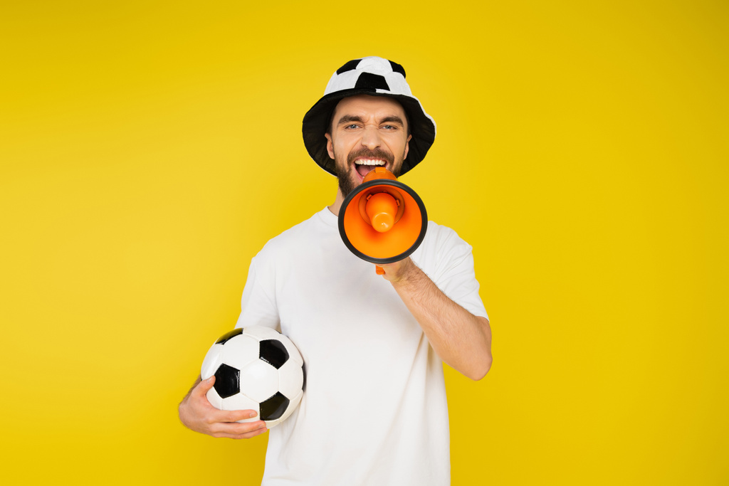 wstrząśnięty człowiek w kapeluszu kibica piłki nożnej stojąc z piłką nożną i krzycząc w głośniku izolowanym na żółto - Zdjęcie, obraz