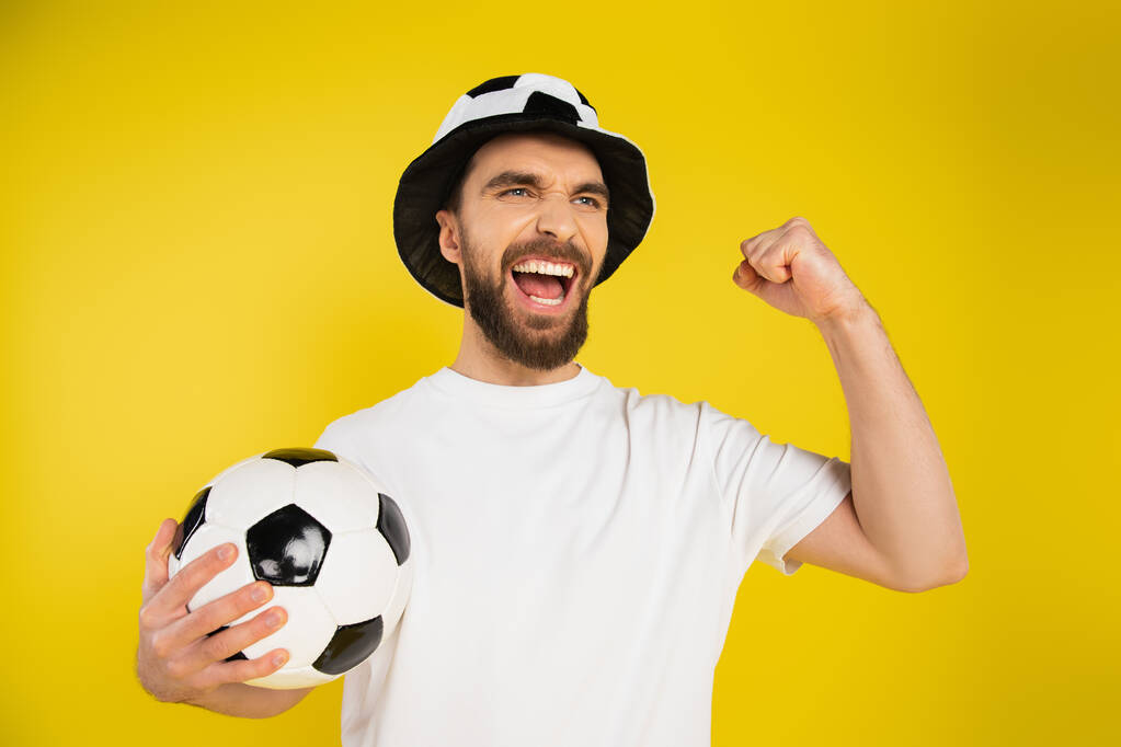 gritando ventilador de fútbol en sombrero sosteniendo pelota de fútbol y mostrando el gesto de éxito aislado en amarillo - Foto, Imagen