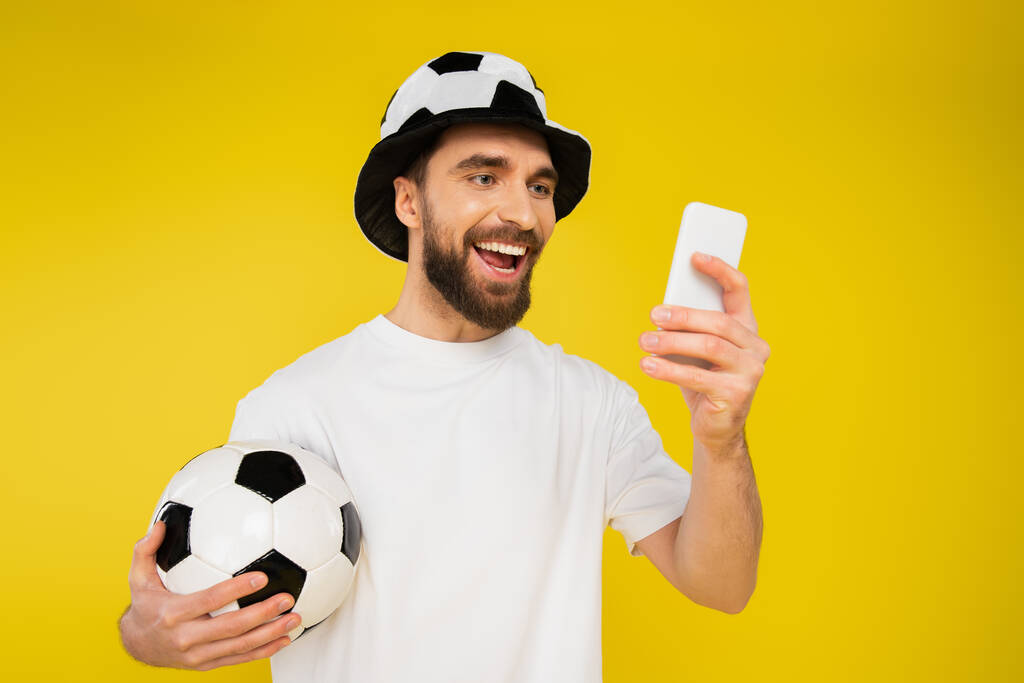 heureux fan de sport dans chapeau regarder match de football sur smartphone isolé sur jaune - Photo, image
