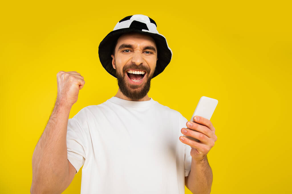 χαρούμενος οπαδός του ποδοσφαίρου στο καπέλο δείχνει χειρονομία θριάμβου και κρατώντας κινητό τηλέφωνο απομονωμένο στο κίτρινο - Φωτογραφία, εικόνα