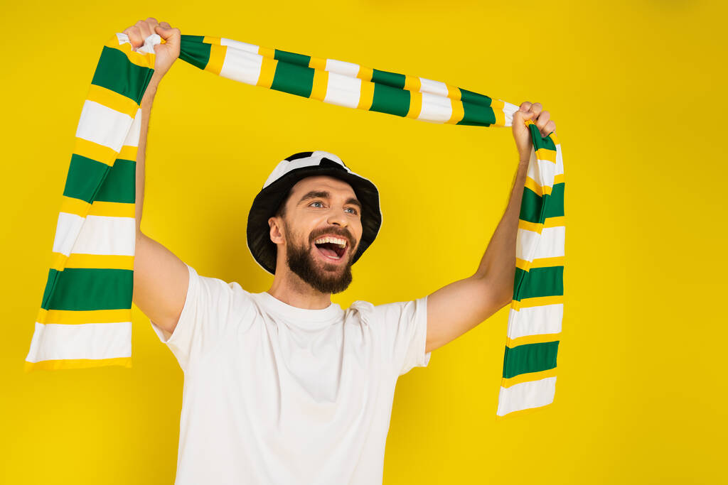 задоволений спортивний фанат в капелюсі кричить, тримаючи смугастий шарф в піднятих руках ізольовані на жовтому
 - Фото, зображення
