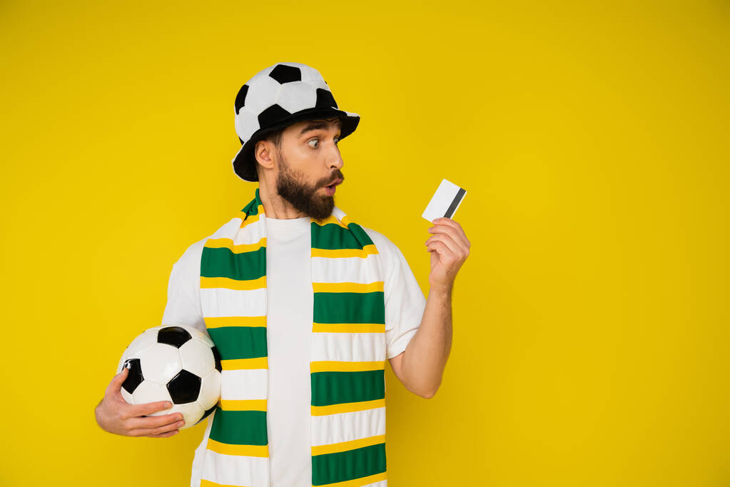 Erstaunter Mann mit Fußballfan-Mütze und gestreiftem Schal, der Fußballball in der Hand hält und die Kreditkarte isoliert auf gelb betrachtet - Foto, Bild