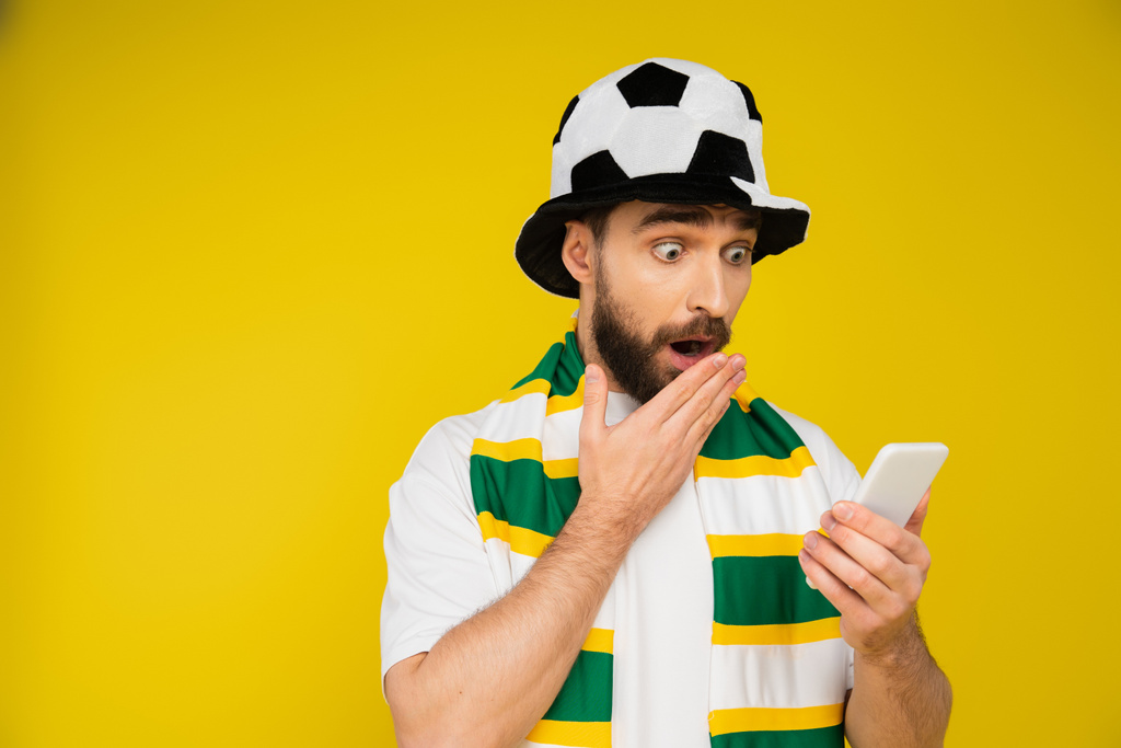 hombre sorprendido en el sombrero del ventilador de fútbol que cubre la boca abierta con la mano mientras mira el teléfono inteligente aislado en amarillo - Foto, Imagen