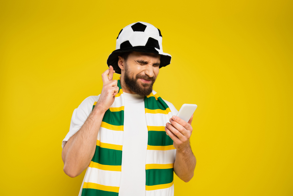 gespannen voetbalventilator met gesloten ogen met gekruiste vingers en mobiele telefoon geïsoleerd op geel - Foto, afbeelding