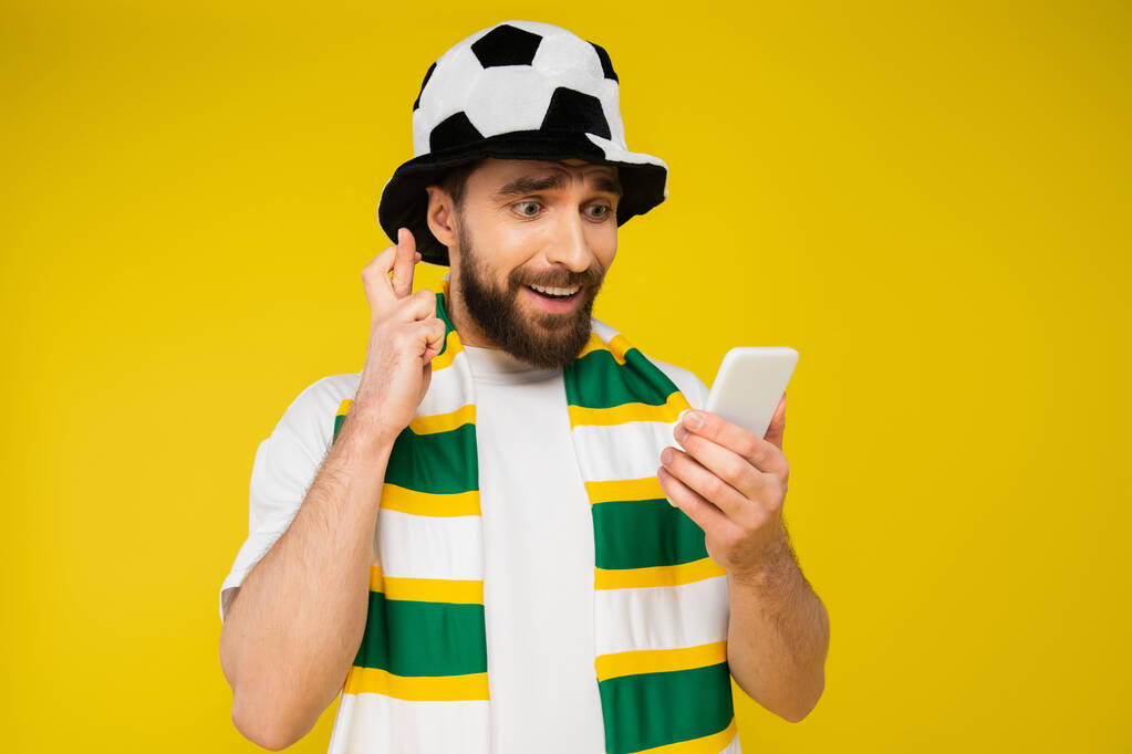 feszült férfi foci fan kalap nézi okostelefon és gazdaság keresztbe ujjak izolált sárga - Fotó, kép