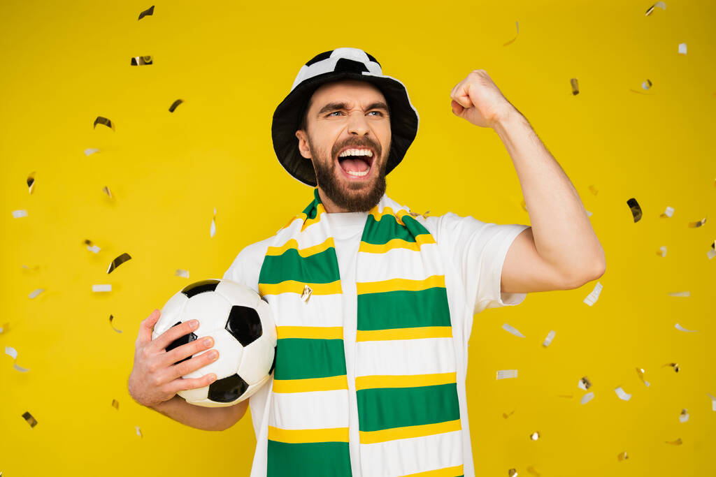 サッカーボールの叫びと黄色の背景に落ちたコンフェッティの下で勝利のジェスチャーを示すと興奮したスポーツファン - 写真・画像