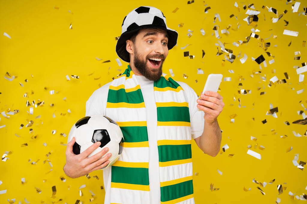 zdumiony i szczęśliwy fan sportu z piłką nożną oglądanie meczu piłki nożnej na smartfonie w pobliżu confetti na żółty  - Zdjęcie, obraz