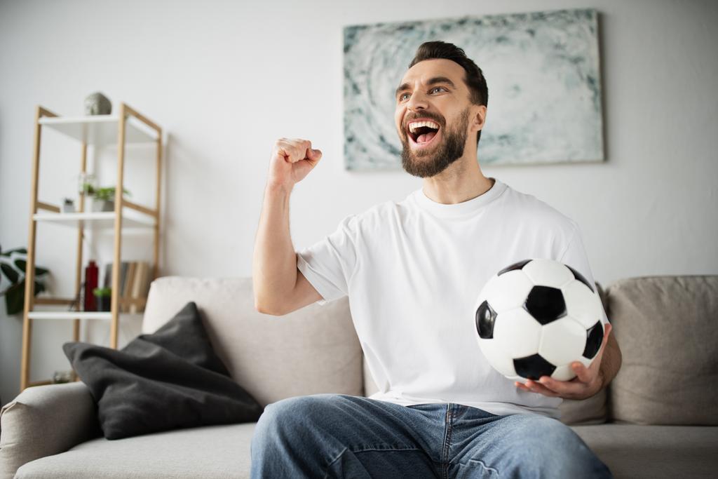 ενθουσιασμένος οπαδός του ποδοσφαίρου κρατώντας μπάλα και δείχνει χειρονομία θριάμβου, ενώ βλέποντας αγώνα στο σαλόνι στο σπίτι - Φωτογραφία, εικόνα