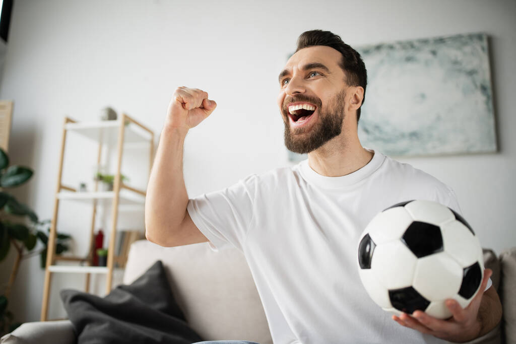shouting bearded man holding soccer ball and showing win gesture while watching football match at home - Photo, Image
