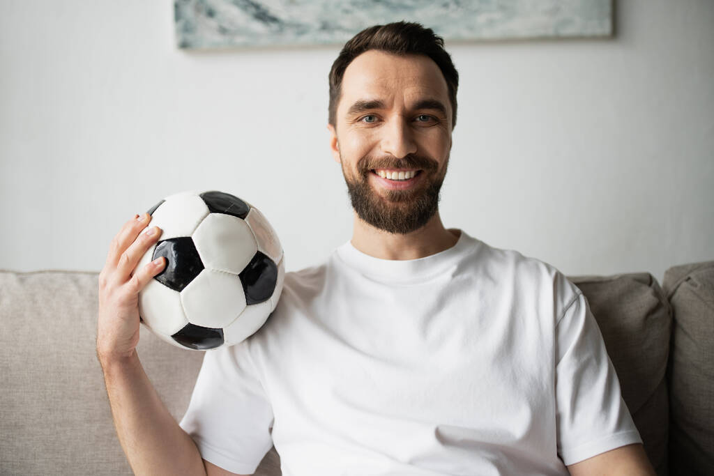 torcedor de futebol alegre e barbudo segurando bola de futebol e sorrindo para a câmera - Foto, Imagem