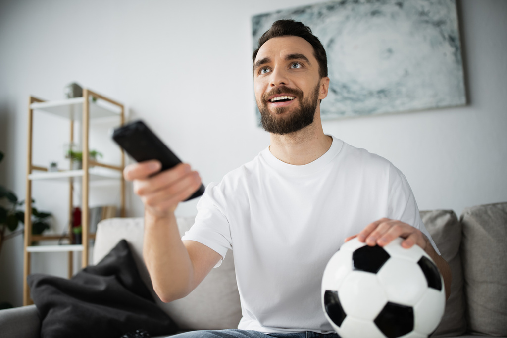 szczęśliwy człowiek trzyma piłkę nożną i pilot podczas oglądania mistrzostw w domu - Zdjęcie, obraz