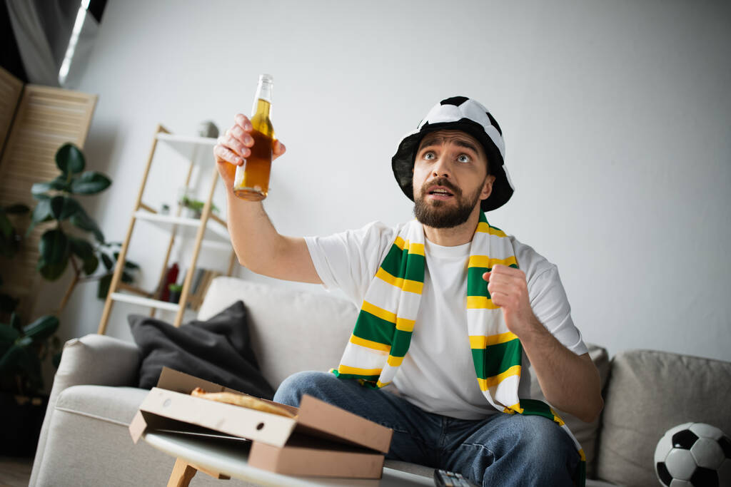 スポーツファンの帽子とスカーフに身を包んだ男がチャンピオンシップを見ながらビールを持っている  - 写真・画像
