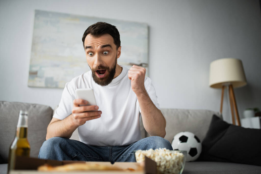 excited man checking football score on smartphone near bottle of beer and tasty food on blurred foreground  - Photo, Image