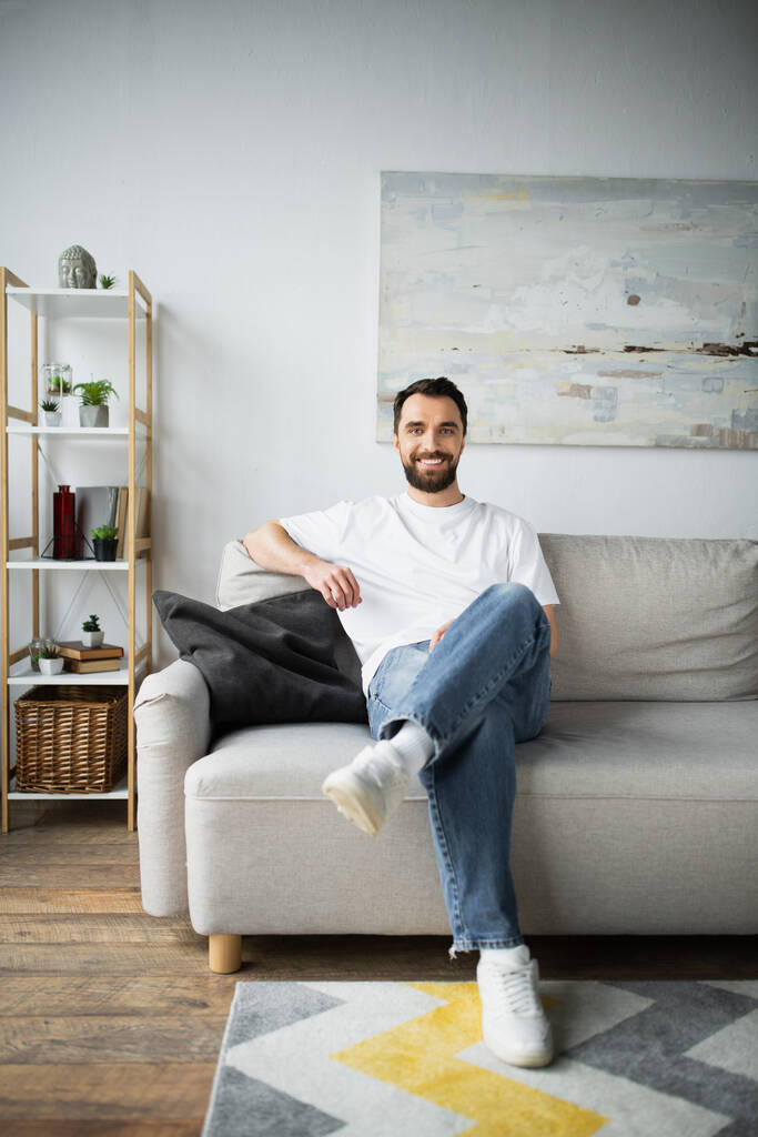 幸せな髭を生やした男でジーンズを着た男がソファの上に座り  - 写真・画像