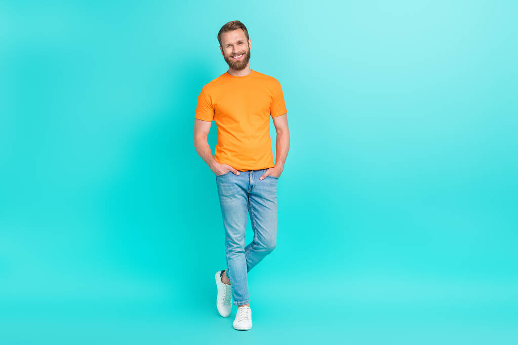 Полный размер фото хорошего настроения красивый мужчина с мужской бородой одет оранжевые руки футболки в карманы изолированы на бирюзовом фоне. - Фото, изображение