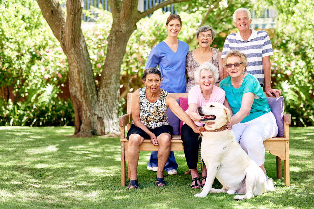 Żyją z witalnością. Portret grupy uśmiechniętych seniorów i pielęgniarki na zewnątrz z labradorem - Zdjęcie, obraz