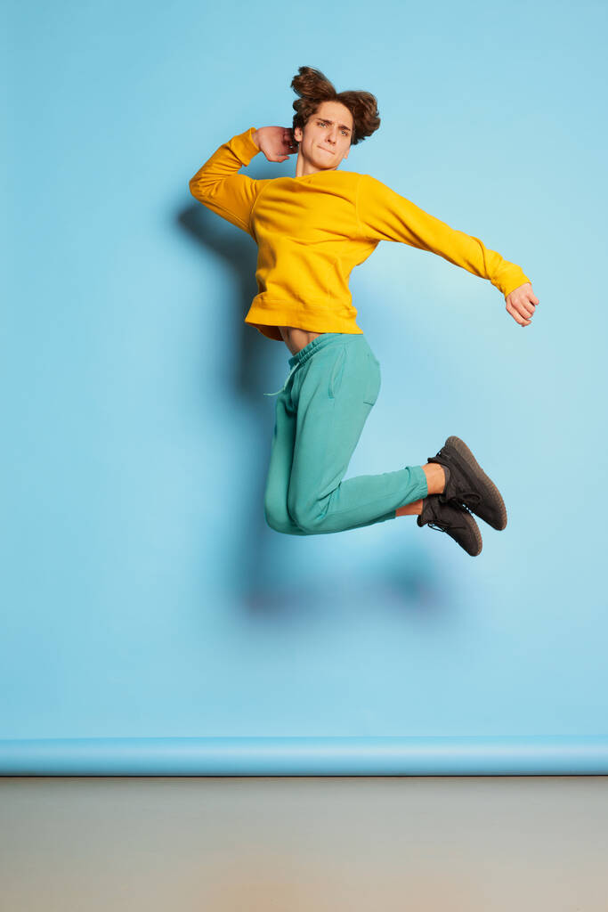 Portrait de jeune homme aux cheveux bouclés posant en vêtements lumineux décontractés et sautant isolé sur fond bleu. Concept de culture de la jeunesse, émotions, expression faciale, mode. Espace de copie pour la publicité - Photo, image