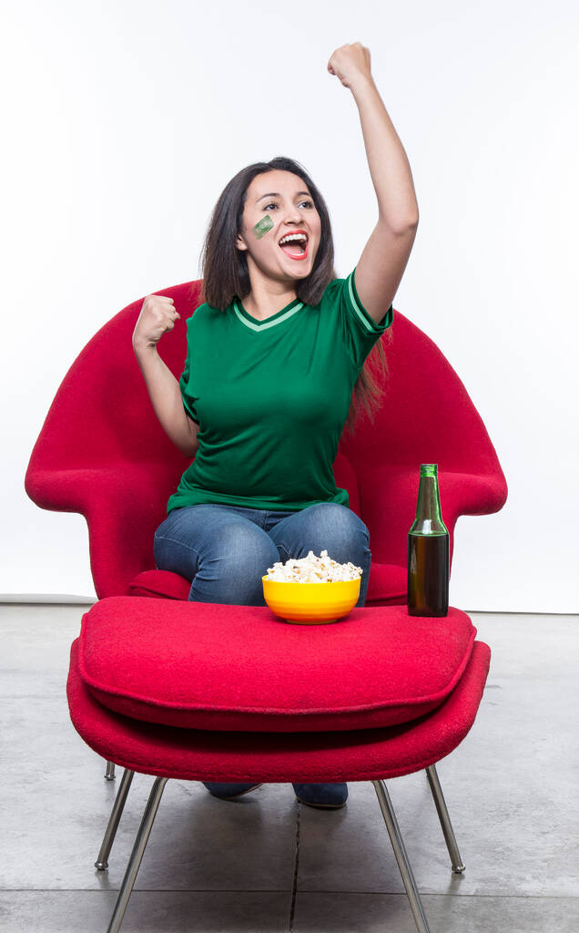 Hermosa joven fan en una camiseta verde y bandera pintada en la cara de la selección nacional de Arabia Saudita celebrando feliz sentado en un sillón rojo con un sándwich y cerveza en un taburete. - Foto, imagen