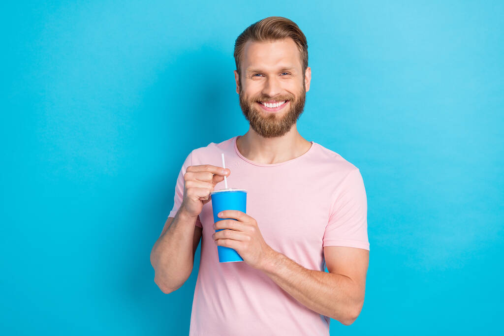 Фото чарівного веселого хлопця, одягненого в рожеву футболку, посміхаючись пити холодний солом'яний напій ізольований синій колір фону
. - Фото, зображення