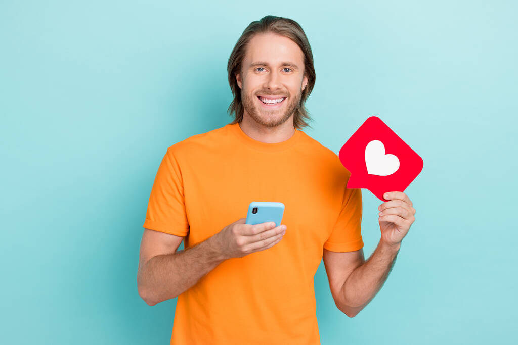 明るいポジティブな男の写真は、現代的なデバイスを入力サインのようなオレンジのTシャツの上昇心臓を身に着けている孤立したティール色の背景. - 写真・画像