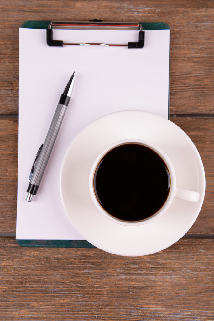 Φλιτζάνι καφέ στο πιατάκι με φύλλο χαρτί και στυλό σε ξύλινο τραπέζι φόντο - Φωτογραφία, εικόνα