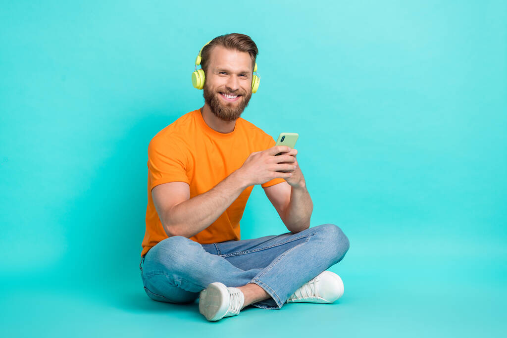 Teljes hosszúságú fotó jóképű optimista fickó visel narancssárga póló ül hallgatni podcast fülhallgató elszigetelt kékeszöld színű háttér. - Fotó, kép