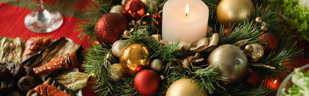 Wianek świąteczny z płonącą świecą i błyszczącymi bombkami w pobliżu grillowanych warzyw na stole z uroczystą kolacją, sztandar - Zdjęcie, obraz