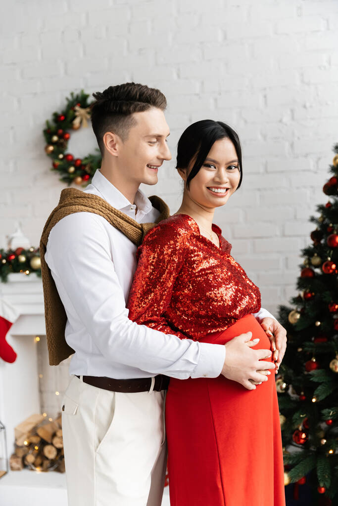 妊娠中のアジアの女性はカメラで笑顔夫は彼女の腹を抱擁しながら彼女の装飾されたリビングルーム - 写真・画像