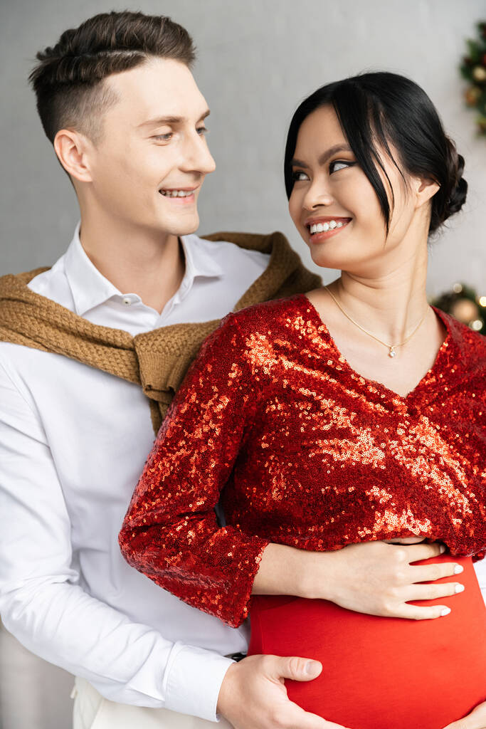 homem feliz e mulher asiática grávida em roupas festivas sorrindo um para o outro - Foto, Imagem