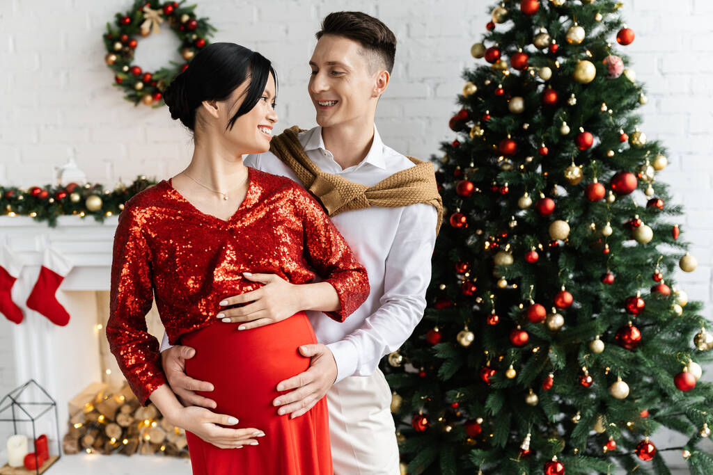 cheerful man and pregnant asian woman in elegant clothes smiling at each other in living room with Christmas decor - Photo, Image