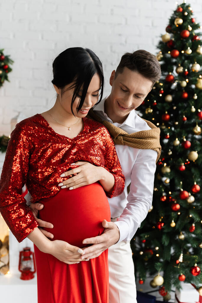 pozitivní muž dotýká břicho těhotné asijské manželky ve svátečním oblečení v blízkosti vánoční strom na rozmazaném pozadí - Fotografie, Obrázek