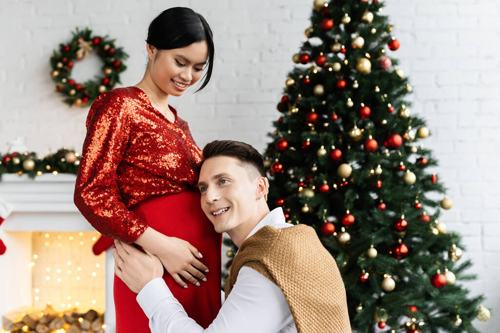 feliz hombre apoyado en la barriga de embarazada y elegante asiático mujer cerca de árbol de Navidad - Foto, Imagen