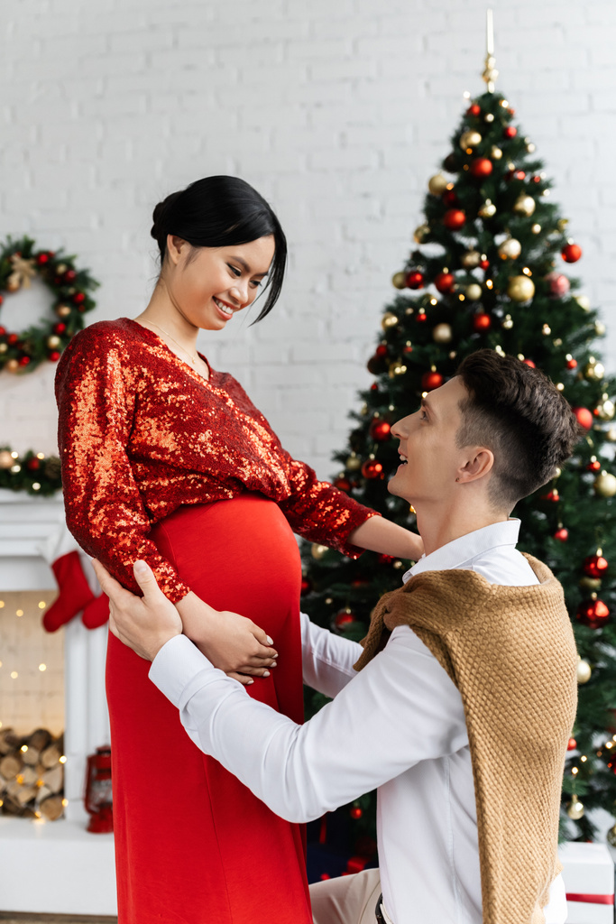 Молодой человек и его беременная азиатская жена в праздничной одежде улыбаются друг другу возле размытой елки - Фото, изображение