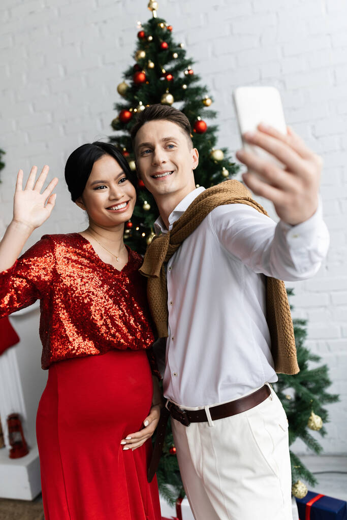 Mutlu bir adam cep telefonuyla hamile Asyalı bir kadının yanında Noel günü video görüşmesi sırasında el sallıyor. - Fotoğraf, Görsel