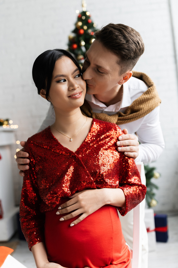 男はクリスマスのお祝いの服で妊娠中のアジアの妻とキス - 写真・画像